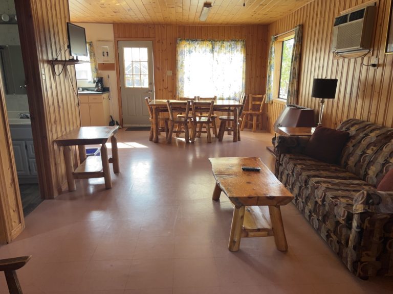 Interior of wood-panelled cottage, with view showing couch, coffee table, air conditioning, and kitchen seating for six