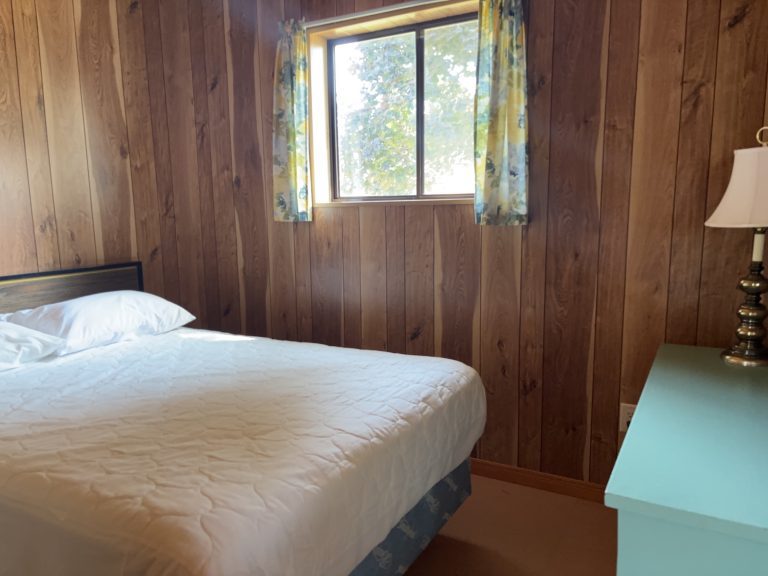 Interior of wood-panelled cottage bedroom, showing bad and window with curtains open