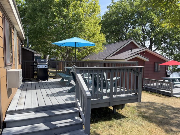 Deck of a wood-panelled cottage, with Muskoka chairs, picnic table, and umbrella