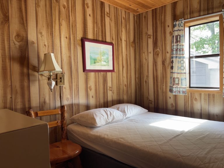 Interior of wood-panelled cottage, with bed by window and picture hanging above it