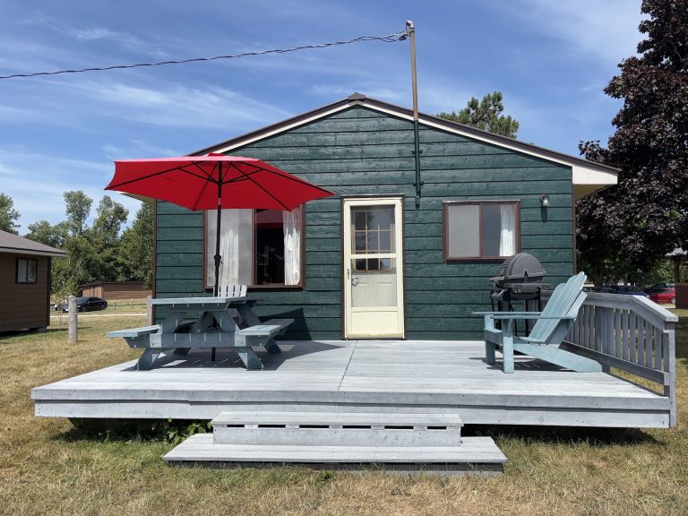 Exterior of green wooden cottage, with barbecue, Muskoka chairs, and red umbrella opened over picnic table