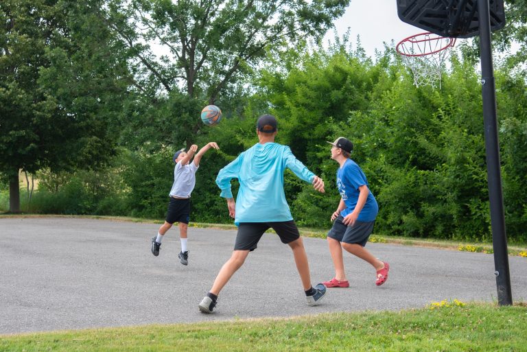 A group of three plays basketball on a paved court, greenery in the backgroudn