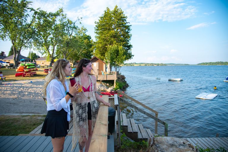 Two people stand by a balcony with drinks and look out towards a lake