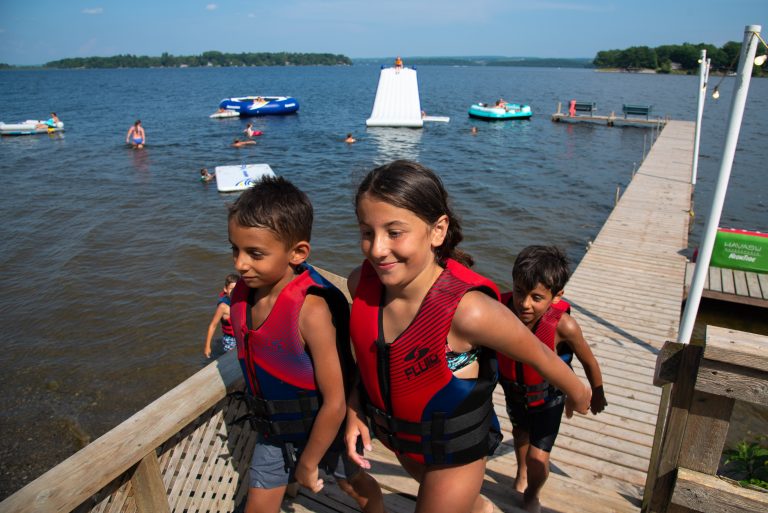A group of kids climbs the stairs up from a dock, with water toys seen in the background