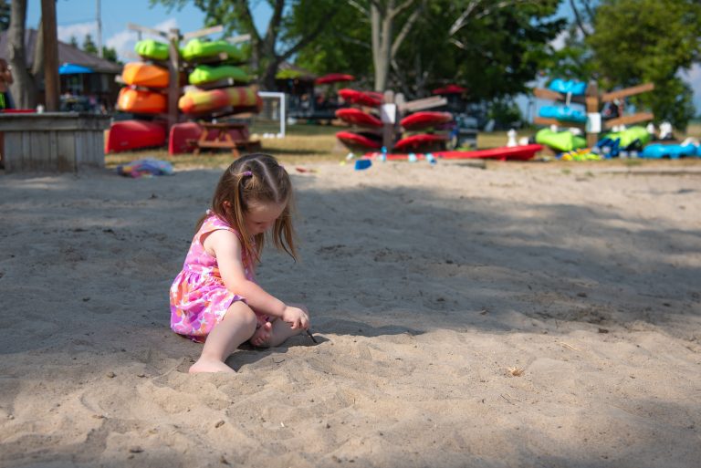 A small child plays with a stick on a sandy beach, kayaks visible in the background