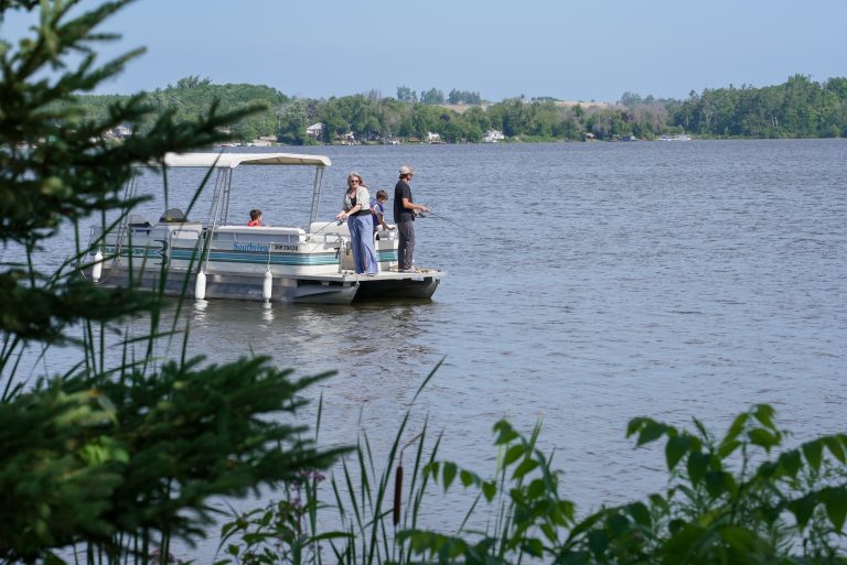 A pontoon boat with people fishing from the back, seen from the shoreline